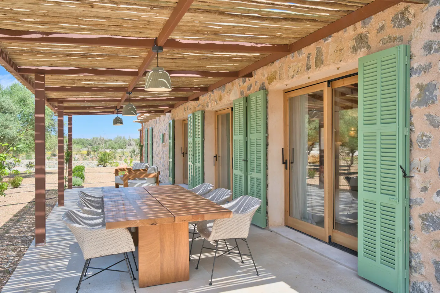 Outdoor patio with a wooden table and chairs under a pergola with green shutters on a stone house.