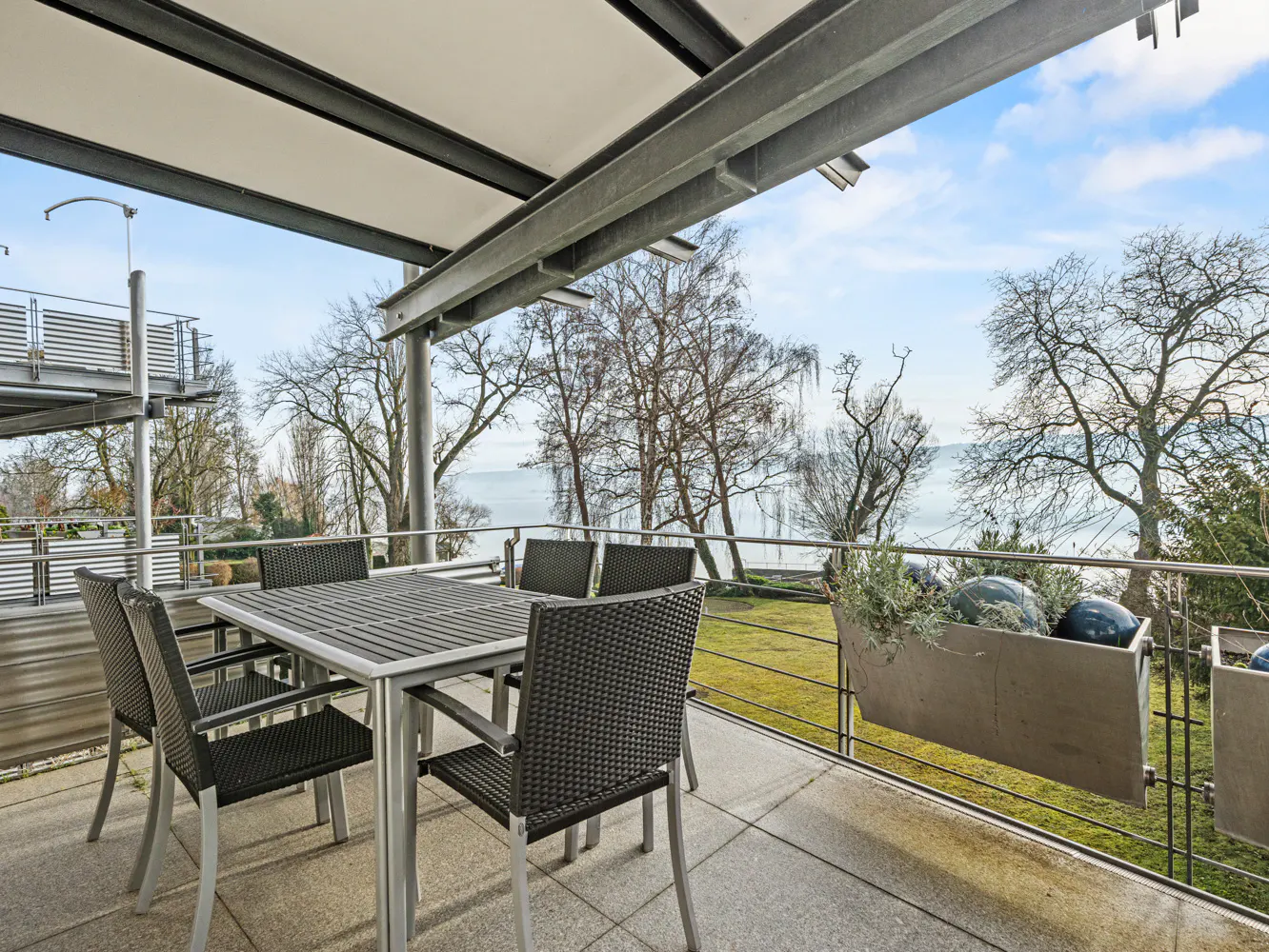 Outdoor patio with a table and chairs overlooking a lake. The patio has a metal frame and a tiled floor.