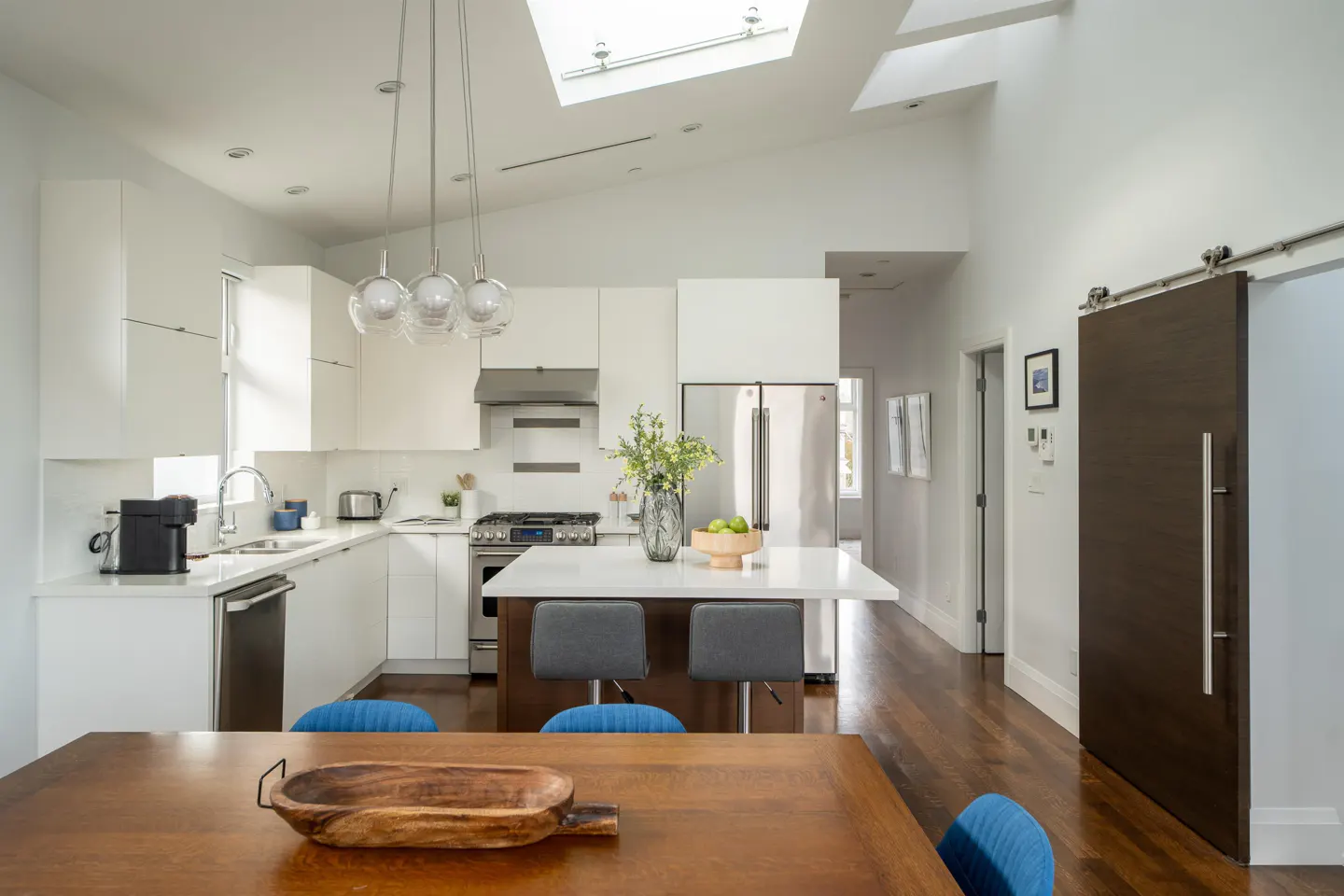 Bright, modern kitchen with white cabinets, stainless steel appliances, and a center island. A wooden dining table is in the foreground.