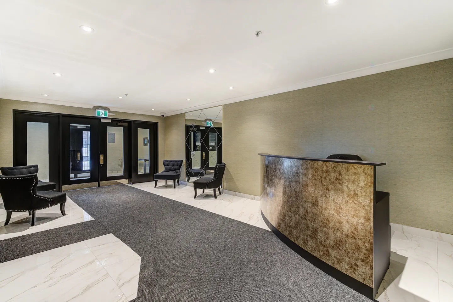 Lobby with a gray carpet, marble floors, and a brown reception desk. Black chairs are near the entrance doors.