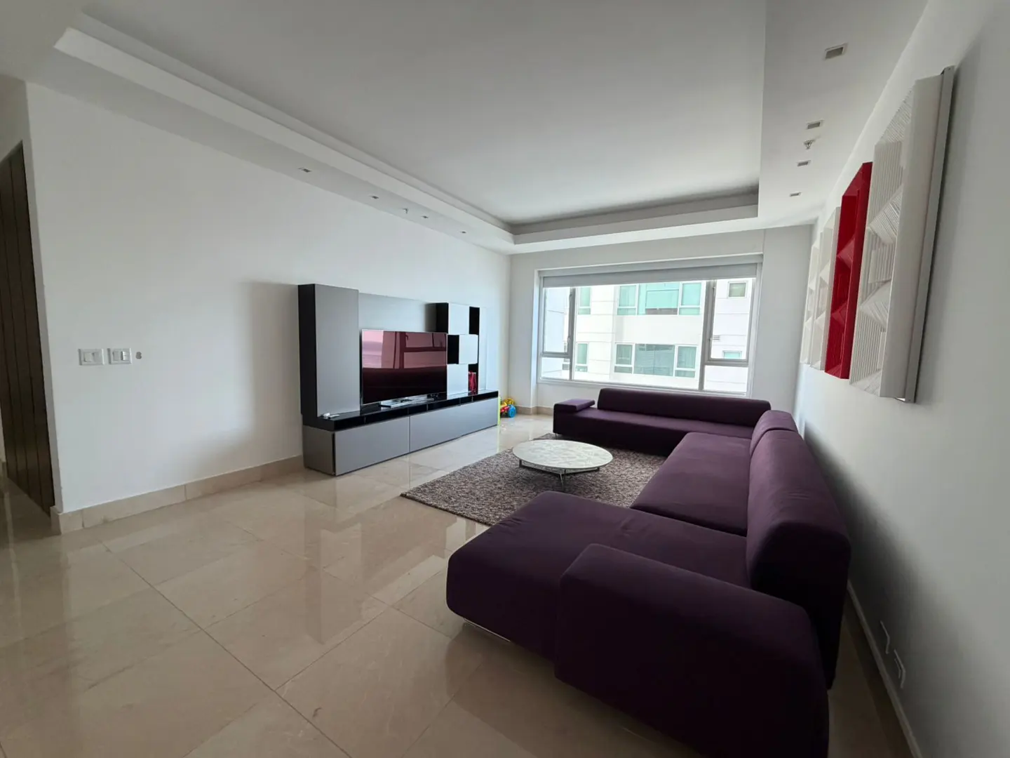 Bright living room with a purple sectional sofa, round marble table, and a gray entertainment center. Large windows offer city views.