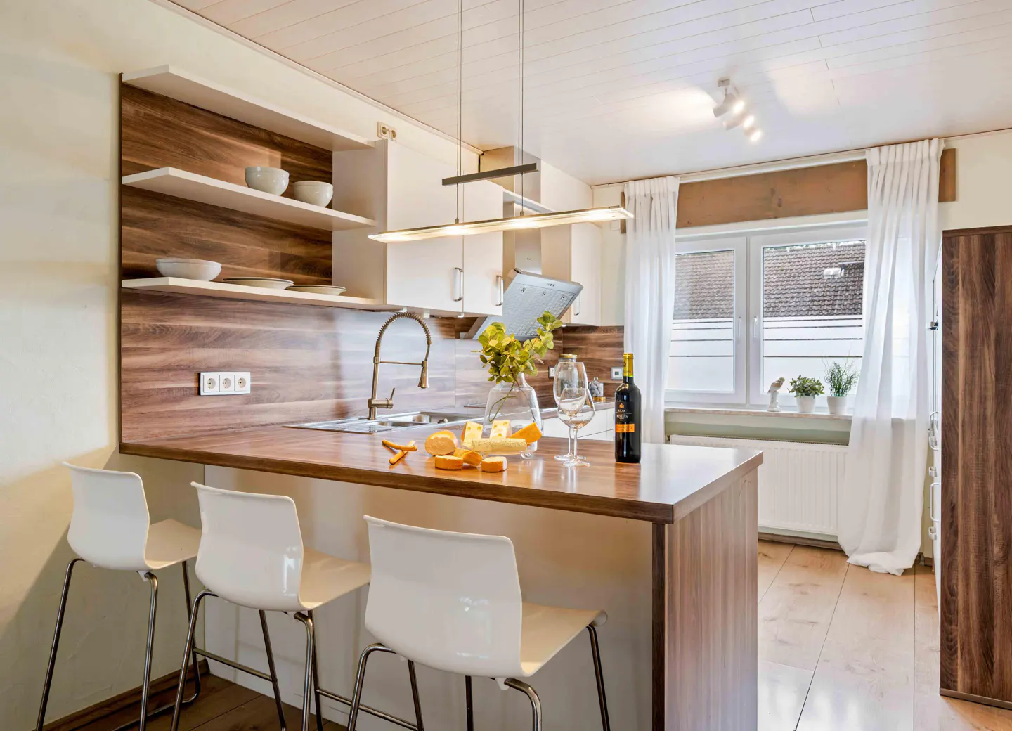 A modern kitchen with white cabinets, wood accents, and a breakfast bar with three white chairs. A window with white curtains is visible.