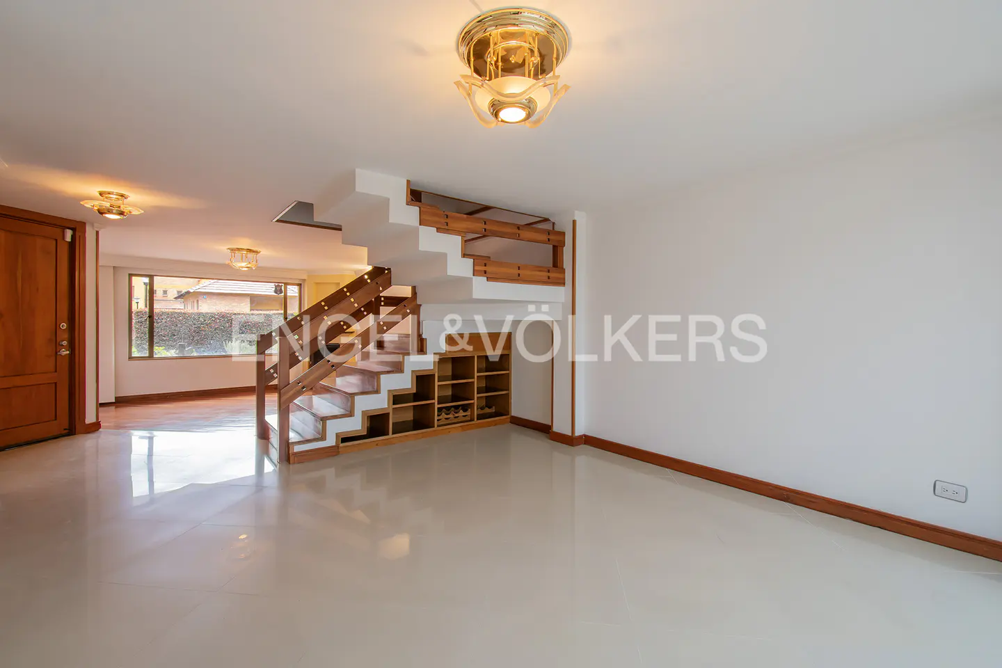Bright, modern living room with white walls and tile floors. A wooden staircase with built-in shelving is visible. A light fixture hangs from the ceiling.