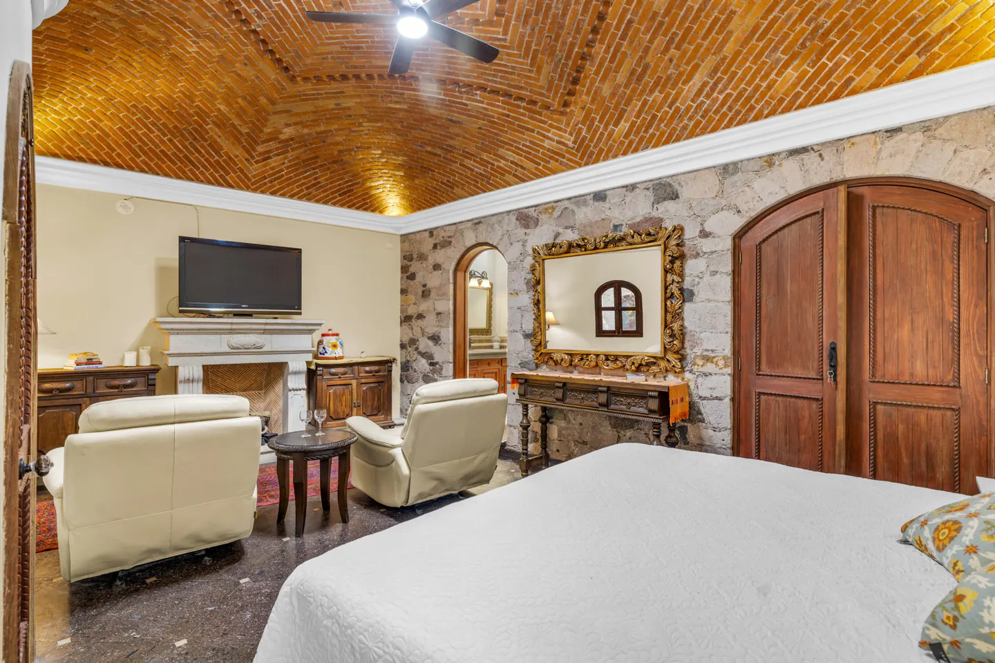 Bedroom with brick ceiling, stone walls, and white bedding. Two cream-colored chairs face a fireplace with a TV above. A large mirror hangs on the wall.