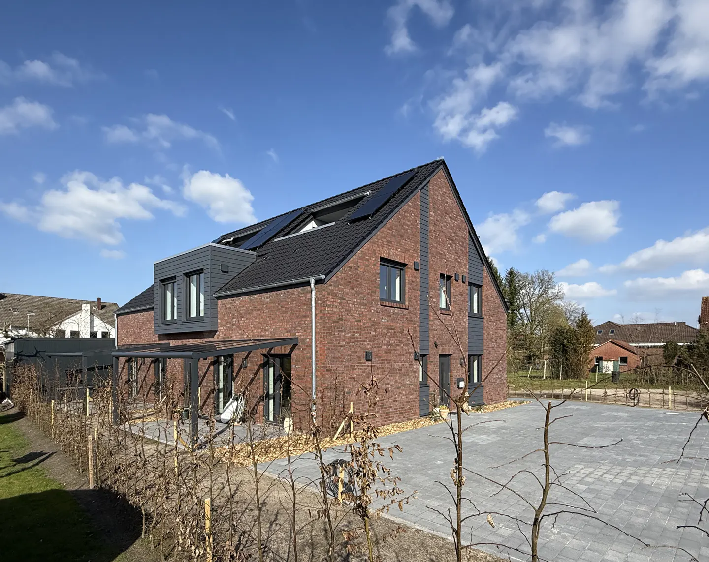 A two-story brick house with a black roof and solar panels, a gray patio cover, and a paved driveway.