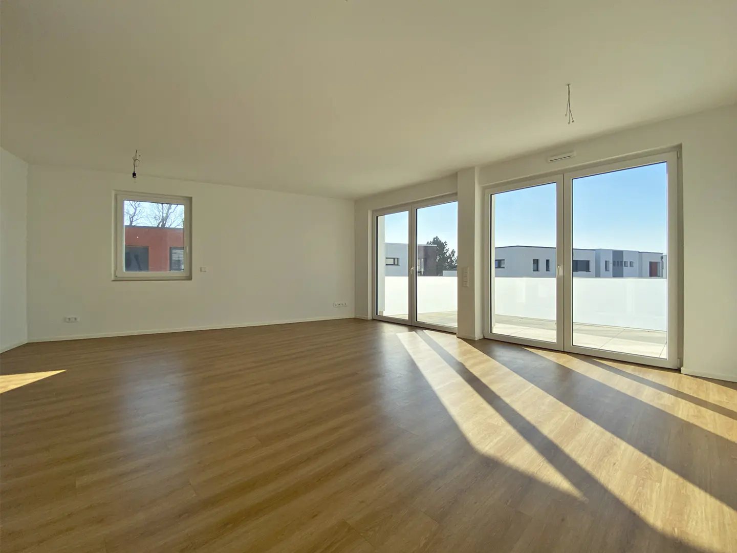Bright, empty room with light wood floors, white walls, and large windows. Sunlight streams in, casting shadows across the floor.