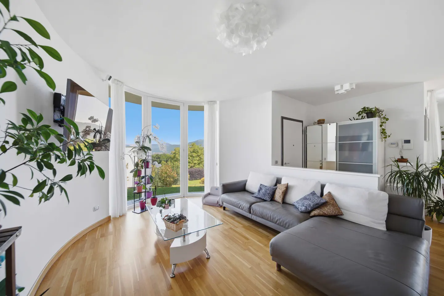 Bright living room with hardwood floors, gray sectional sofa, glass coffee table, and large windows with a view of a green landscape.