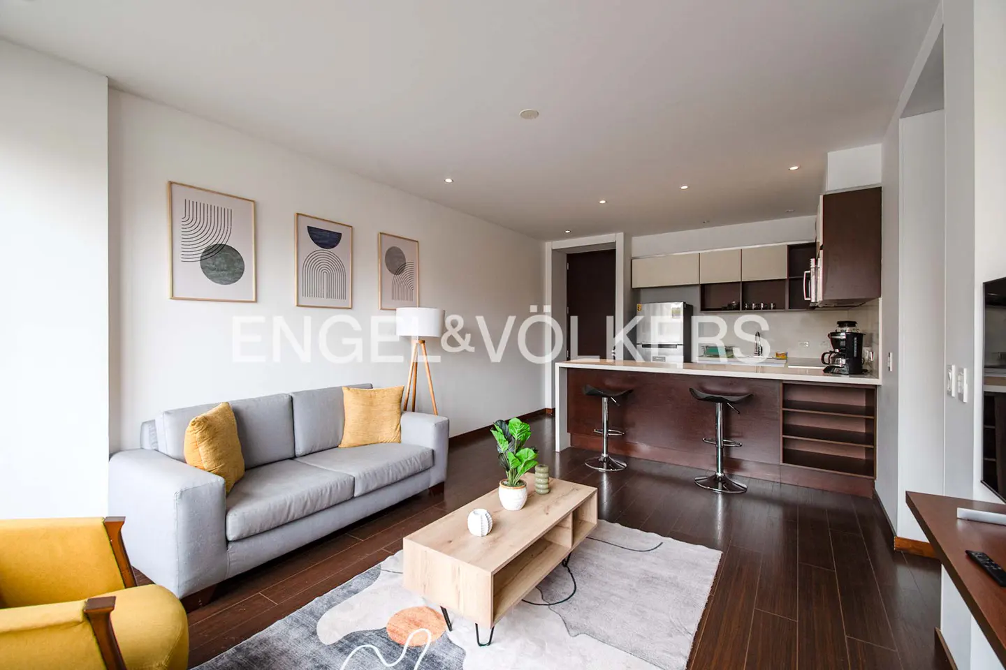 Living room with gray sofa, yellow pillows, wood coffee table, and open kitchen with bar stools. Engel & Völkers logo overlay.