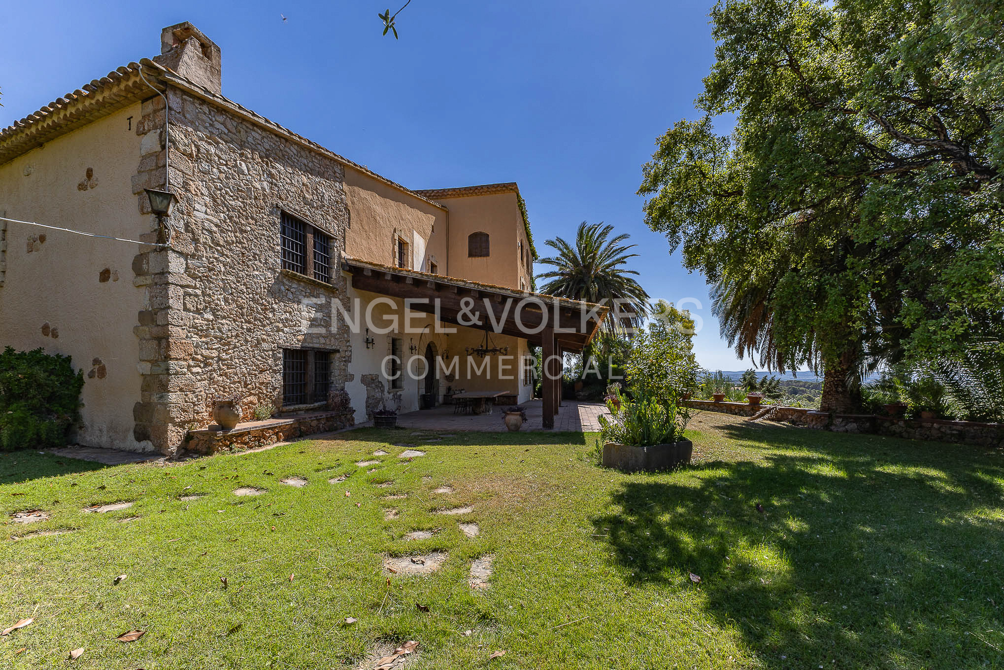A rustic stone and stucco villa with a covered patio, set in a sunny garden with a large tree under a clear blue sky.