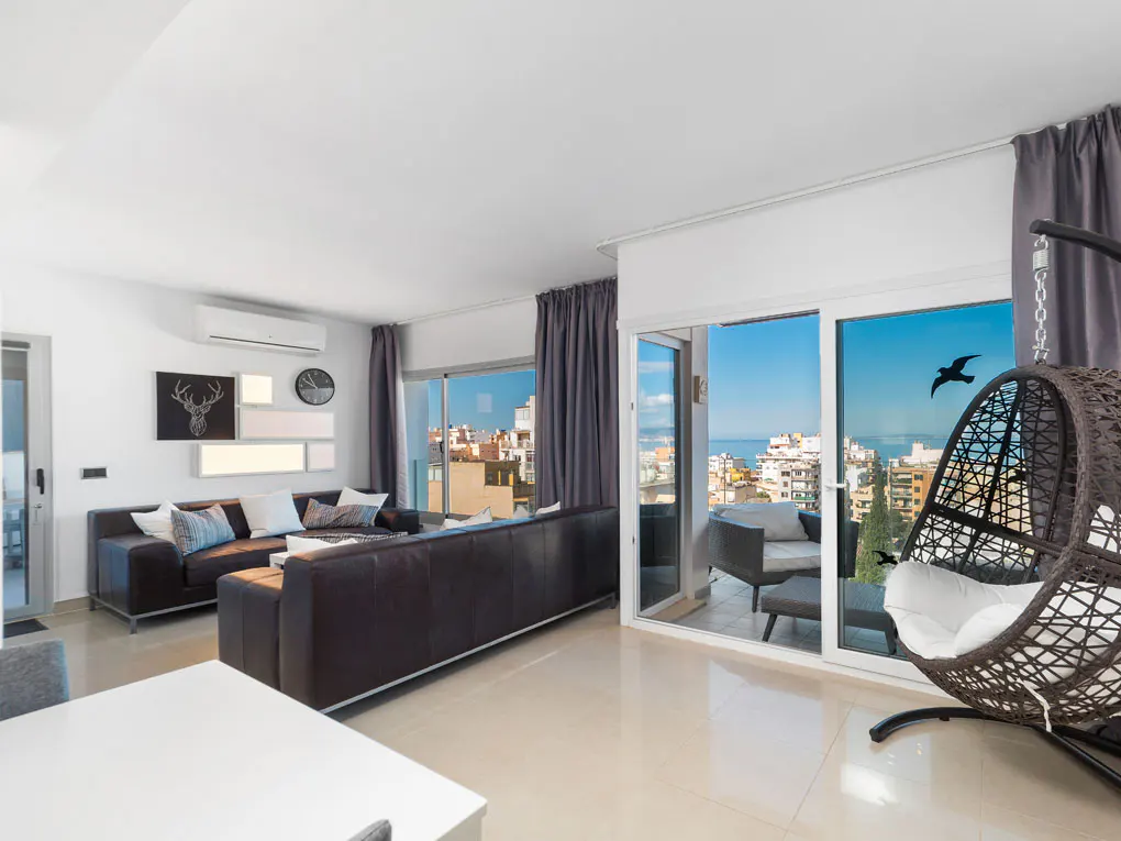 Bright living room with brown leather sofa, white walls, and a balcony with city views. A hanging chair sits near the sliding glass doors.