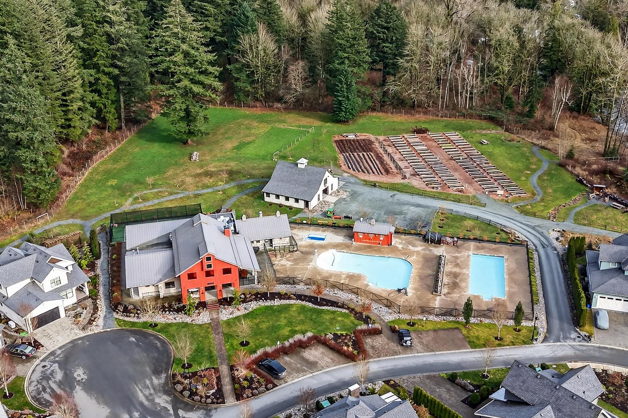 Aerial view of a red farmhouse with two swimming pools, a barn, and garden beds surrounded by a lush forest.