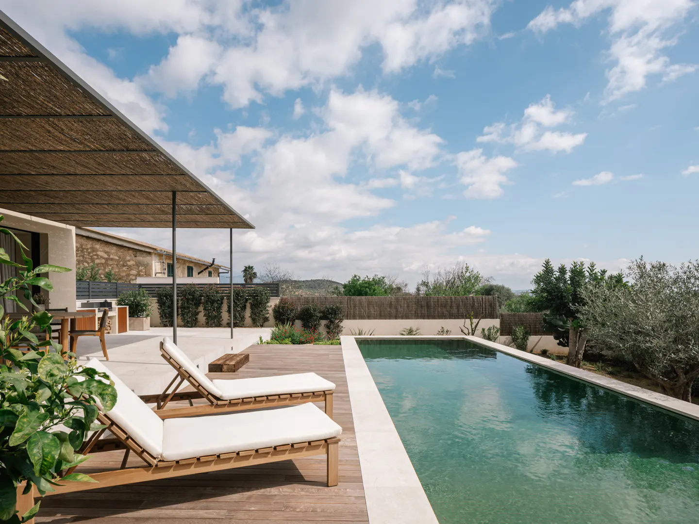 A backyard pool with two lounge chairs on a wooden deck under a thatched roof, with a blue sky and clouds above.