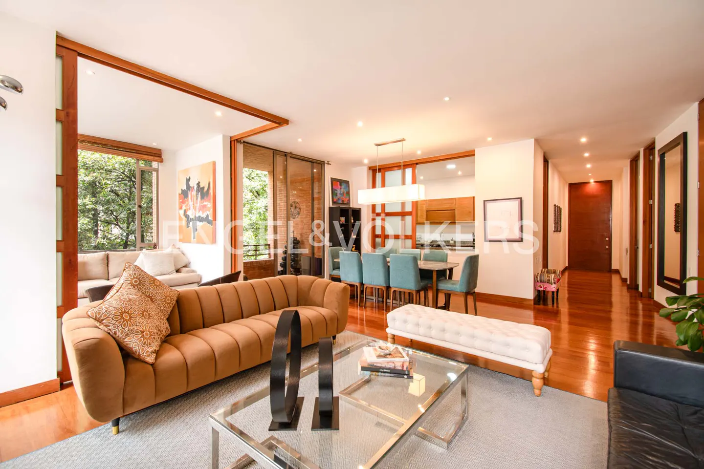 A bright, modern living room with a brown sofa, glass coffee table, and dining area with blue chairs. Large windows offer a view of lush greenery.