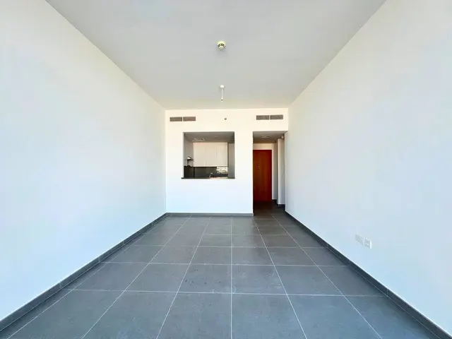 Empty room with white walls and gray tiled floor. A kitchen area is visible through an opening, and a brown door is on the right.
