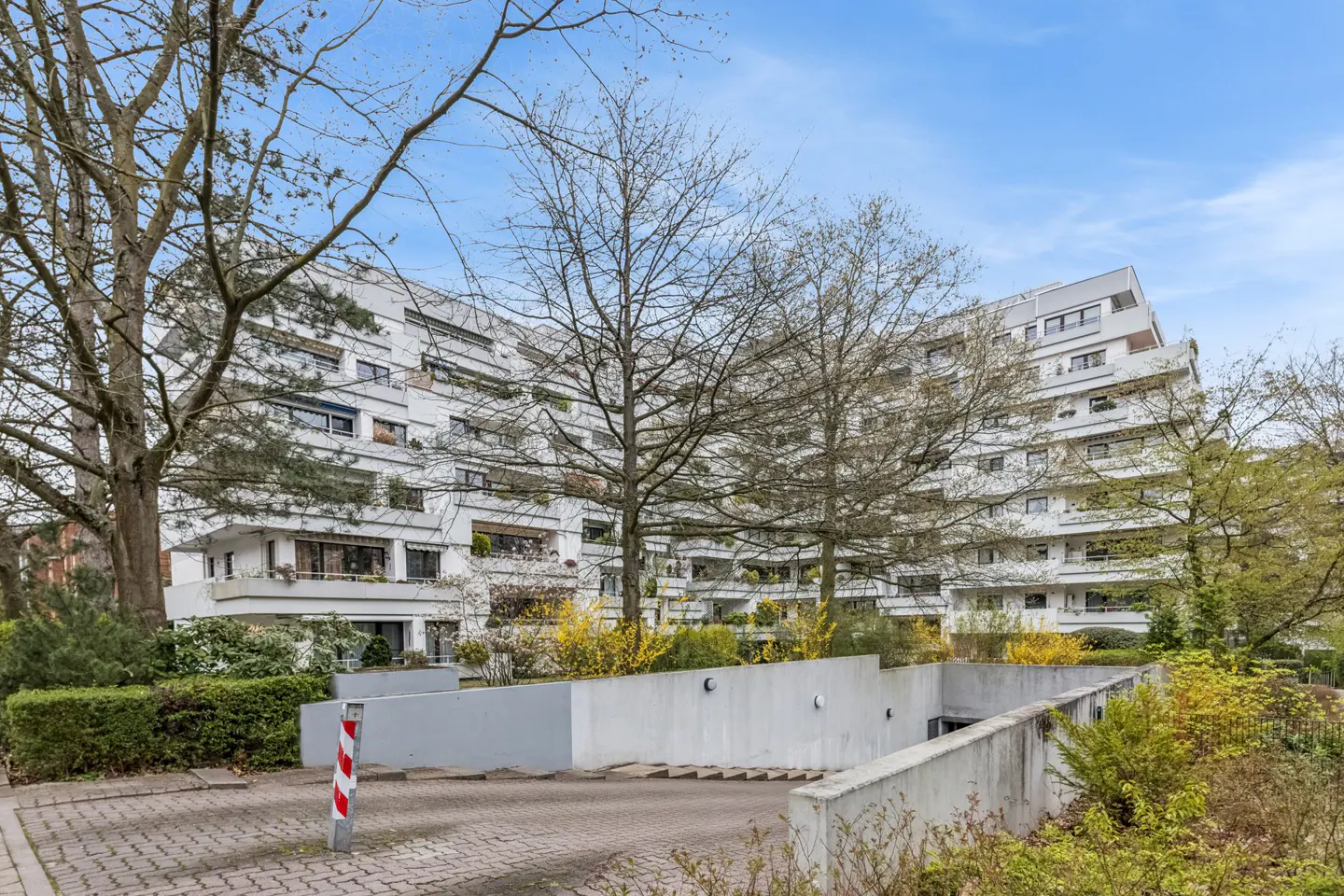 Exterior view of a modern white apartment building with balconies, surrounded by trees and greenery, under a blue sky.