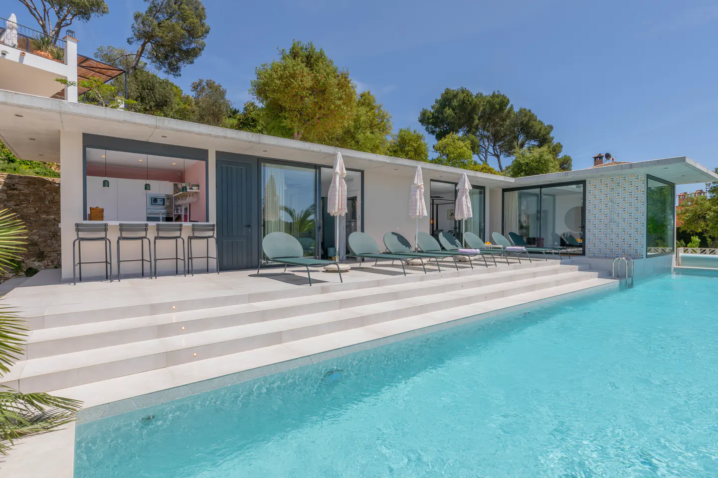 A modern white house with a pool, lounge chairs, and bar stools under a blue sky.