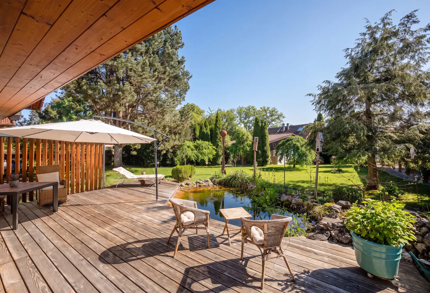 Wooden deck with wicker chairs, a small pond, and a large white umbrella overlooking a lush green backyard garden.