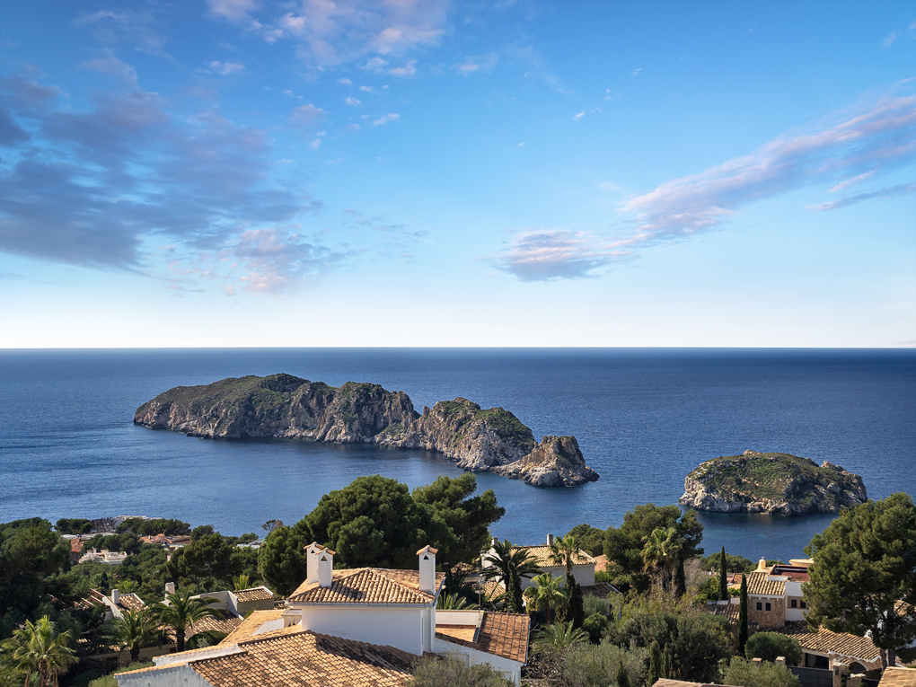 View of the Mediterranean Sea with rocky islands, lush green trees, and white villas with terracotta roofs under blue sky.