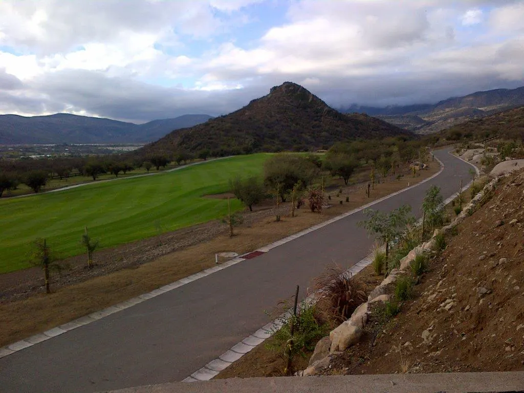 Scenic view of a winding road next to a green golf course, with a mountain backdrop under a cloudy sky.