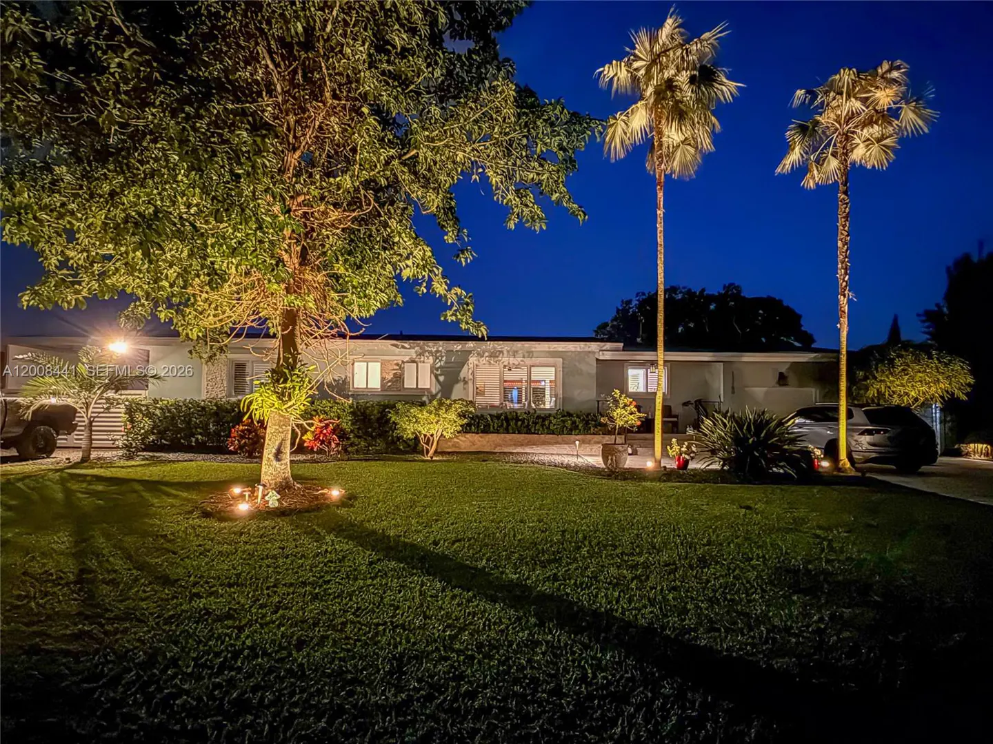Night view of a one-story light gray house with a green lawn, trees, and two tall palm trees illuminated by spotlights. A car is parked in the driveway.