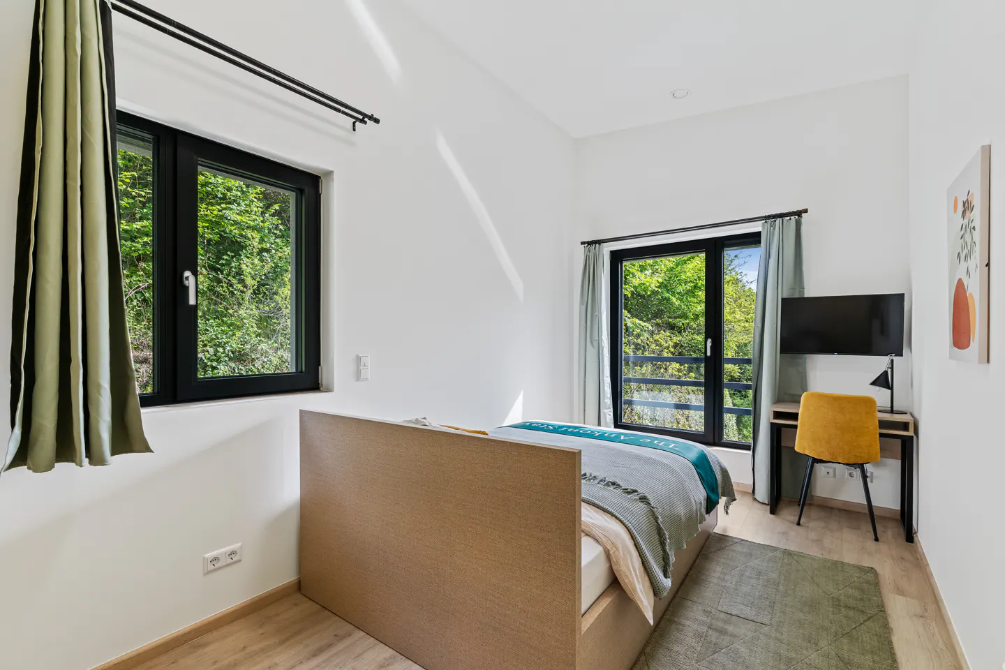 Bright bedroom with white walls, wood floors, and two black-framed windows with green curtains. A bed, desk, yellow chair, and TV are also visible.