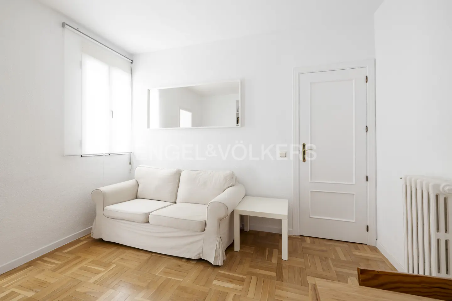 Bright room with white walls, parquet floor, white sofa, small table, window, mirror and white door.