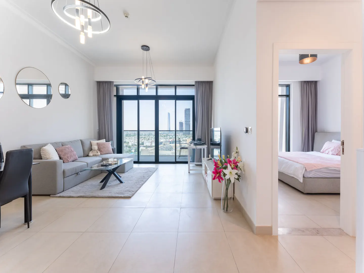 Bright, modern apartment living room with a gray sofa, glass table, and city view through large windows. Bedroom visible through doorway.