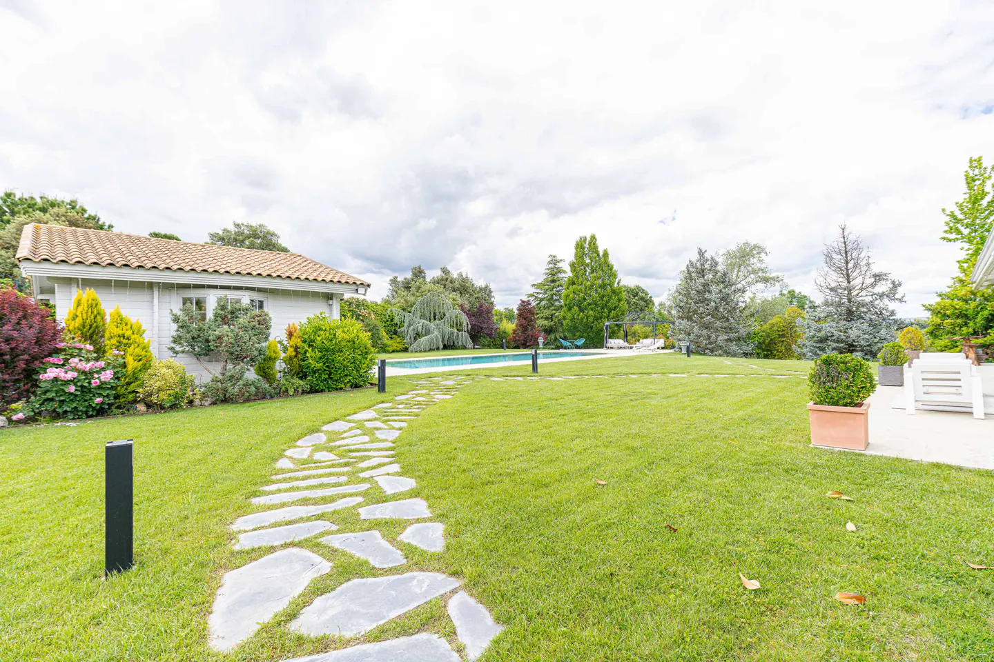 Stone path leads through a green lawn to a pool and white house under a cloudy sky.