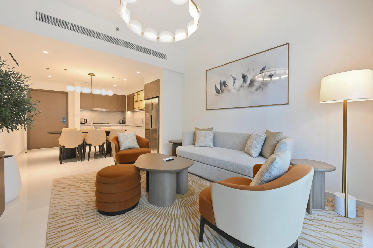 Bright, modern living room with a gray sofa, two orange chairs, and a round coffee table on a patterned rug. Kitchen and dining area visible in the background.