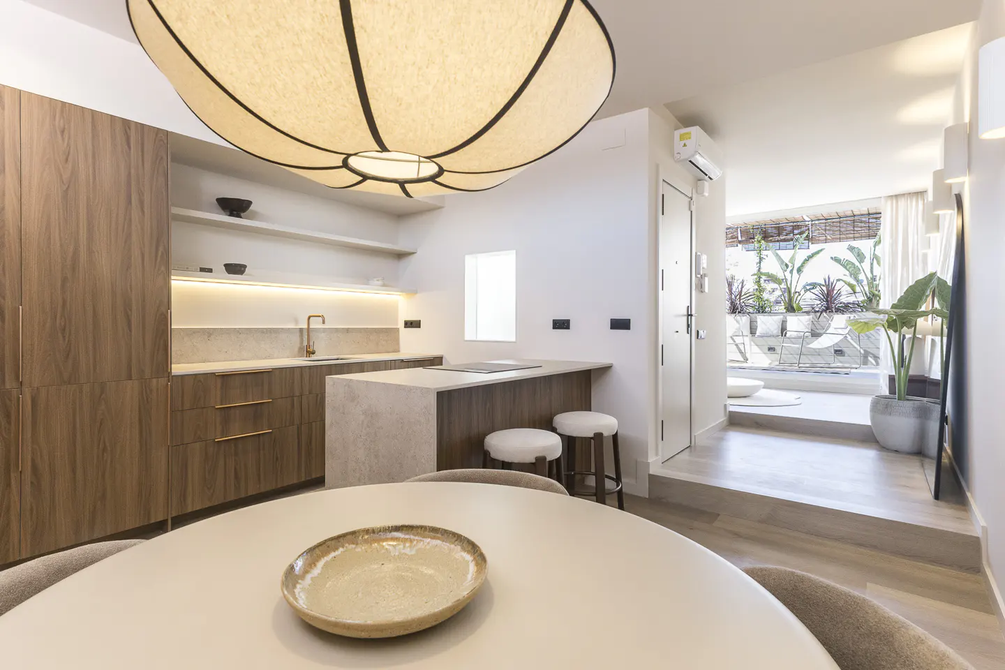 A bright, modern kitchen with wood cabinets, a stone island, and a round dining table. A large, decorative light fixture hangs above. A patio is visible through a doorway.