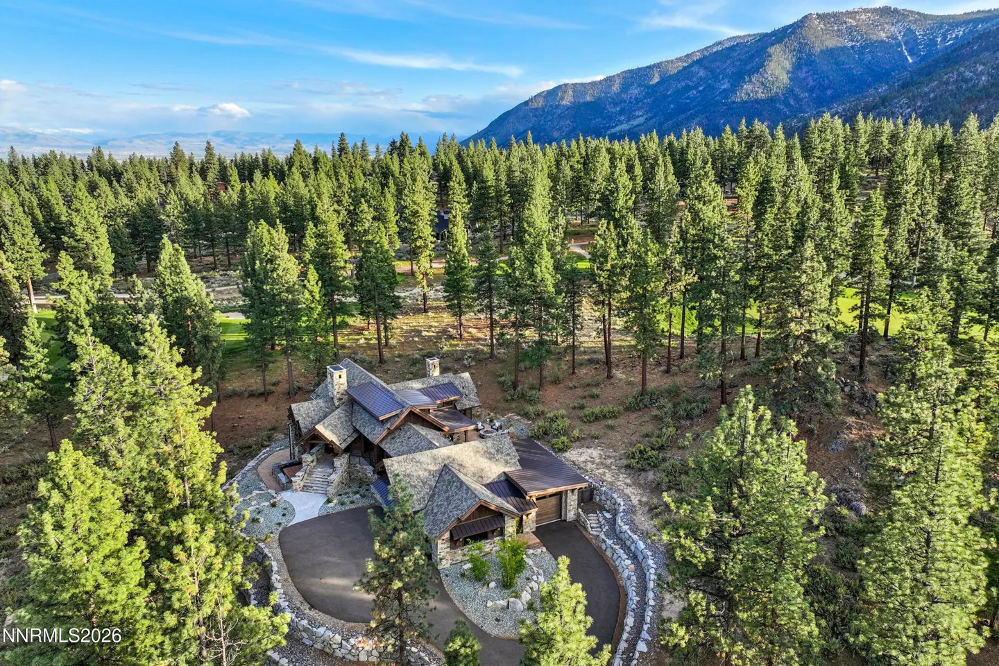 Aerial view of a stone house with solar panels, surrounded by tall green trees and a mountain backdrop under a blue sky.