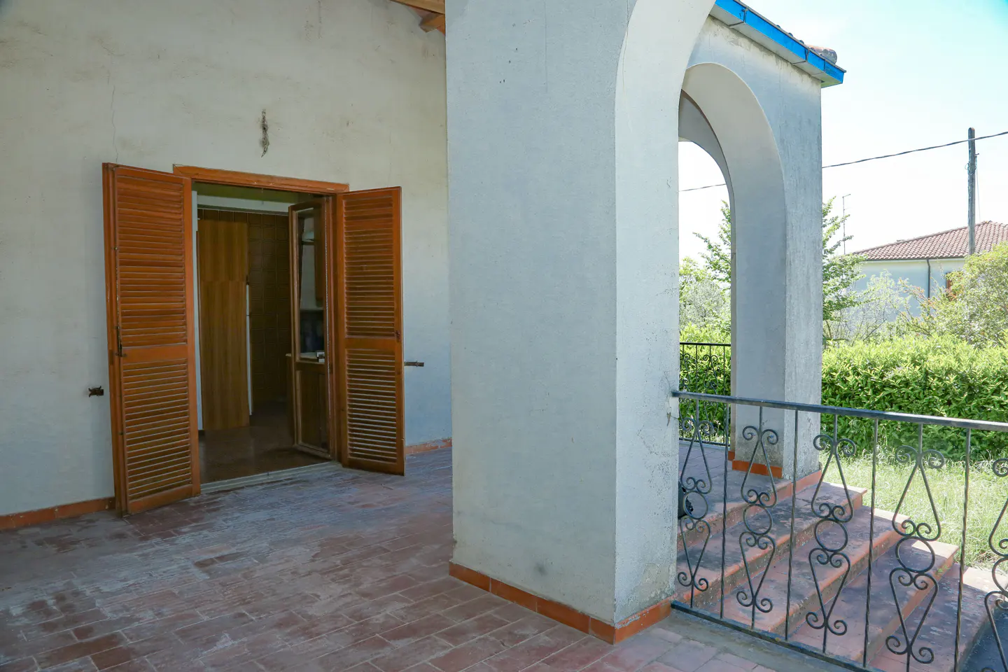 Covered porch with arched columns, terracotta tile floor, and open shuttered doors. Black wrought iron railing overlooks green foliage.