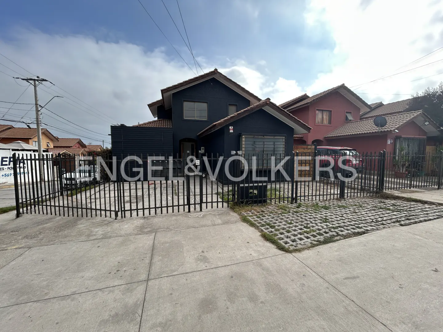 Two-story dark gray house with a tiled roof, enclosed by a black metal fence, viewed from a concrete street.