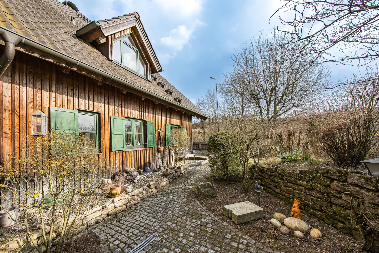 Exterior view of a wood-sided house with green shutters and a stone walkway leading to a garden.