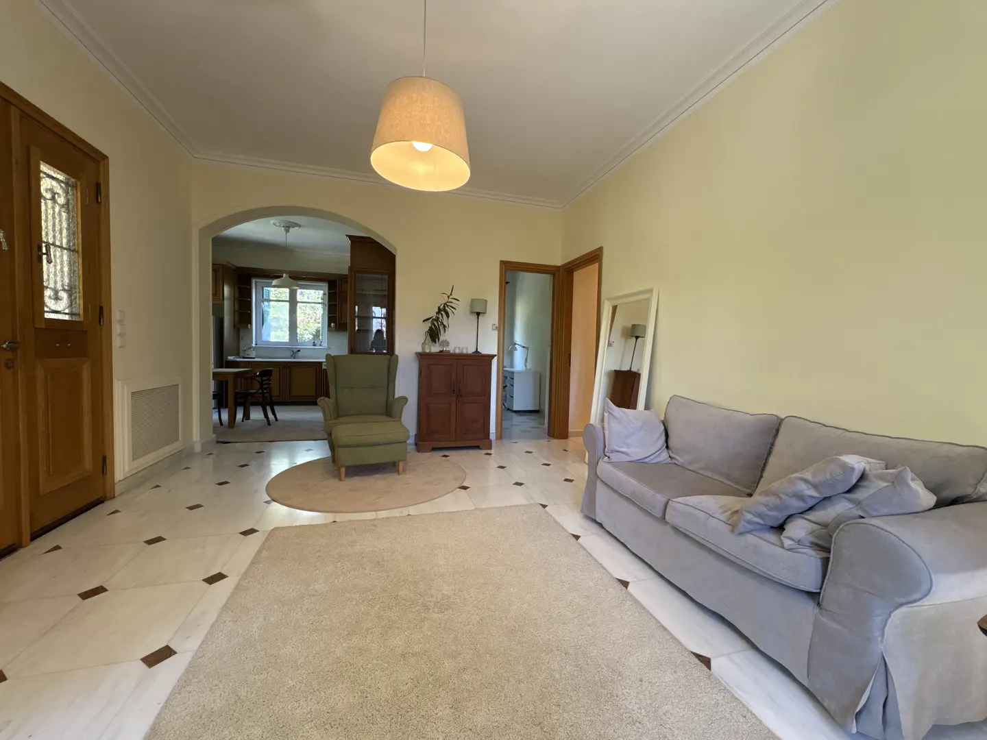 A living room with a gray sofa, green armchair, and marble floors with brown accents. A kitchen is visible through an archway.