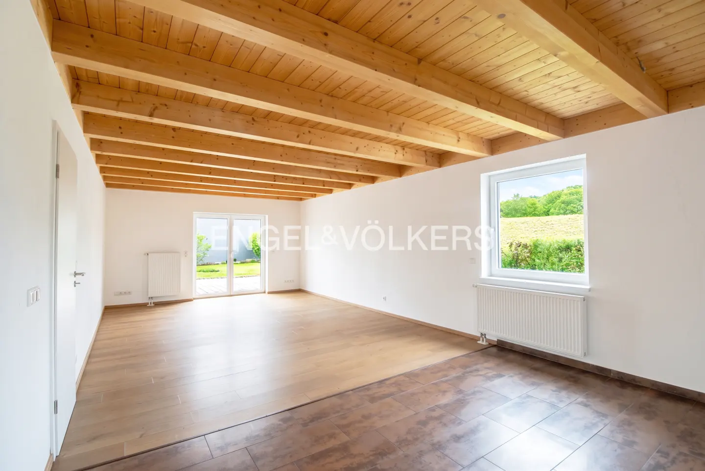 Bright, empty room with wood floors, white walls, exposed wood beam ceiling, and a window overlooking a green field.