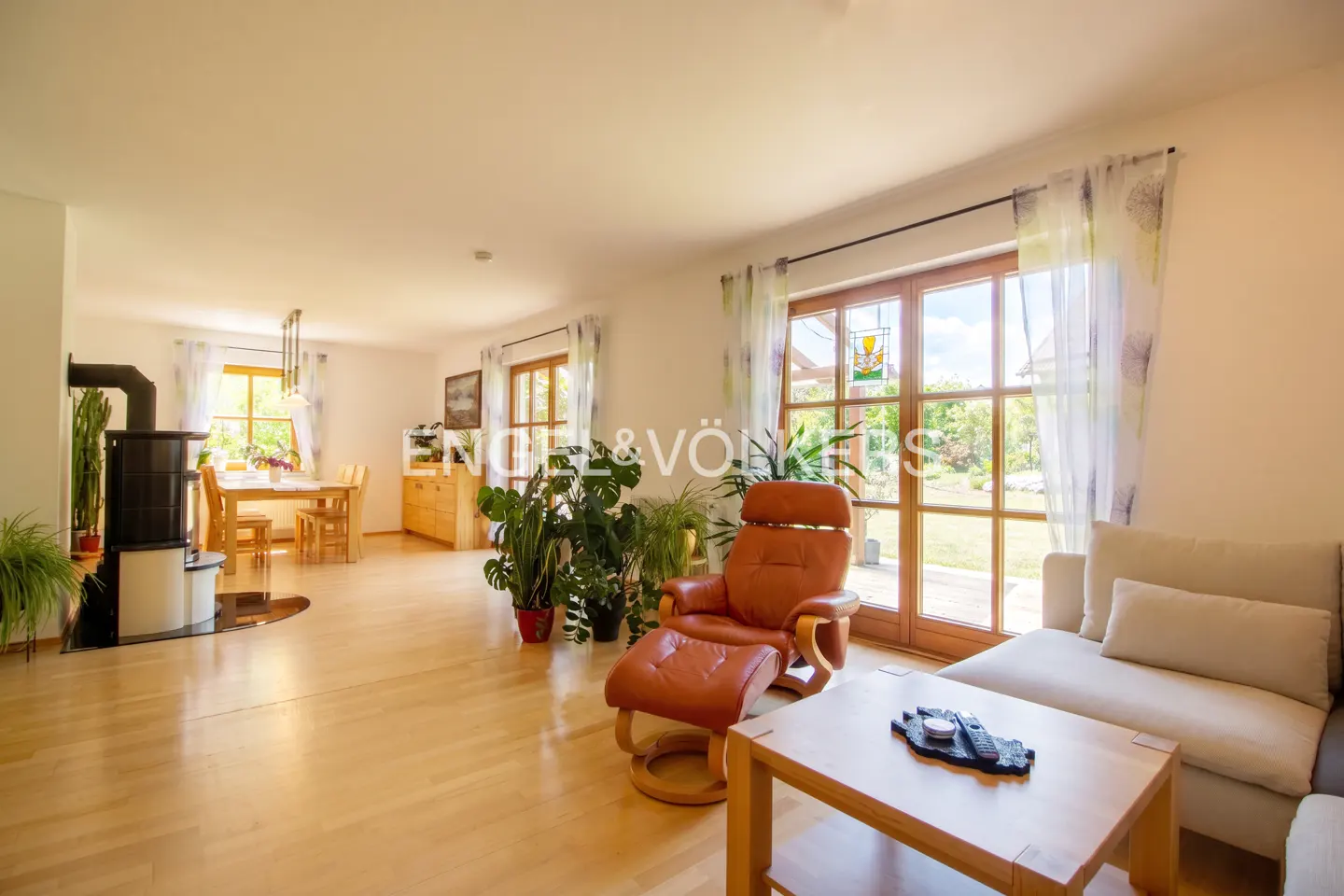 Bright living room with wood floors, a brown leather recliner, and a cream sofa. A wood-burning stove and dining table are visible in the background.