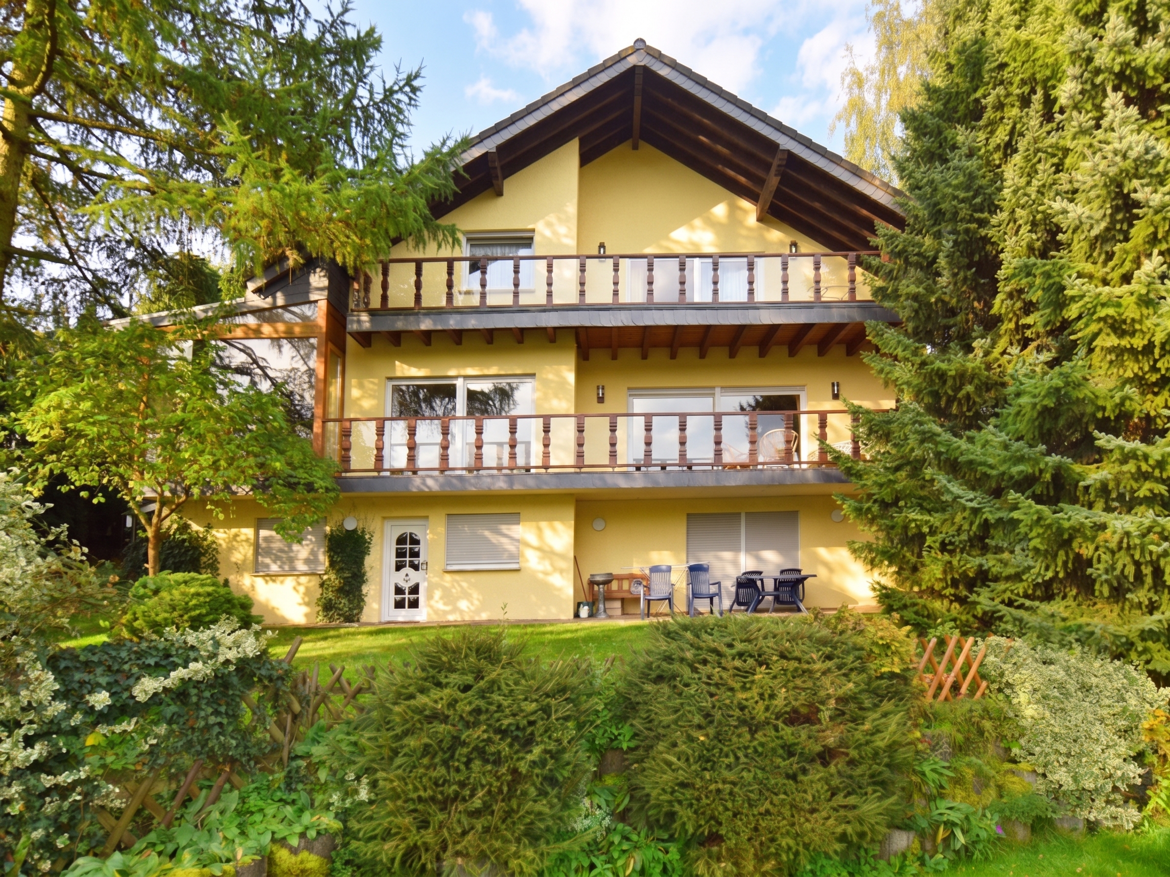 A three-story yellow house with two balconies, surrounded by lush green trees and a garden with patio furniture.
