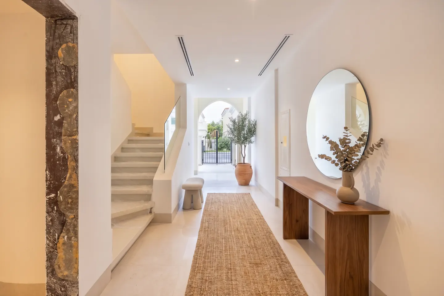 Bright foyer with white walls, beige runner, and stairs. A wooden console table with a round mirror and vase of eucalyptus is on the right.