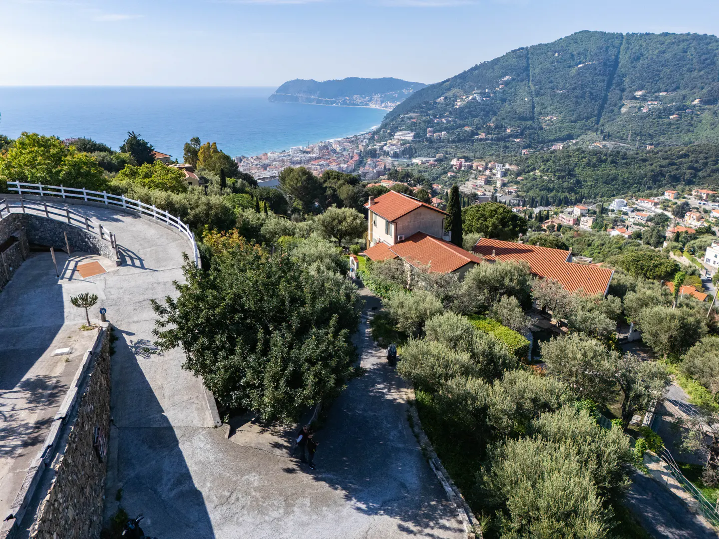 Scenic view of a coastal town with red-roofed houses nestled among green trees, overlooking the blue sea and distant mountains.