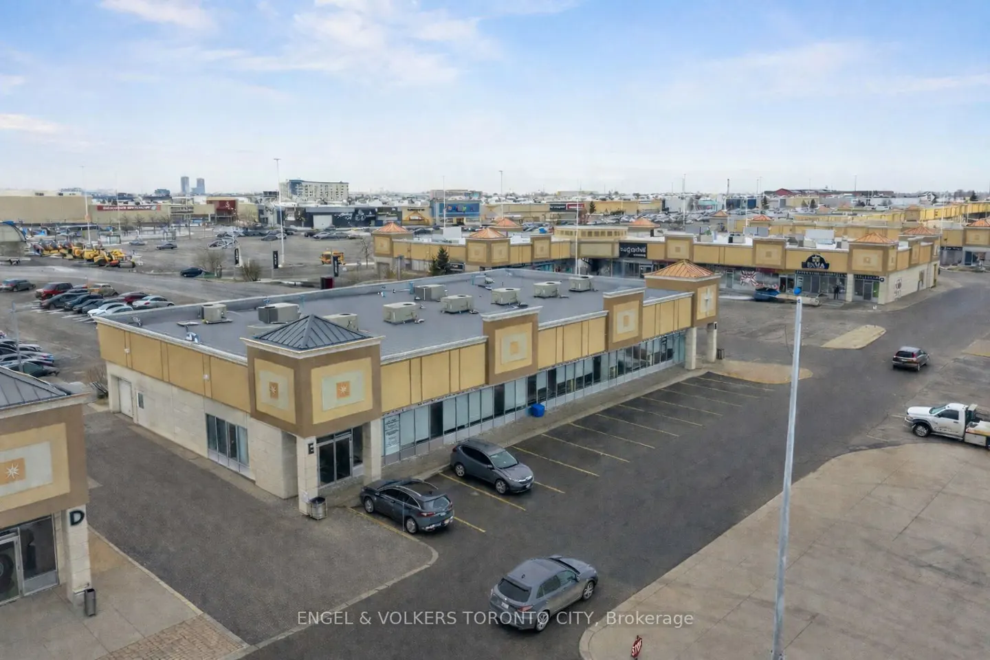 Aerial view of a tan commercial strip mall with a flat roof, parking lot, and surrounding urban landscape.
