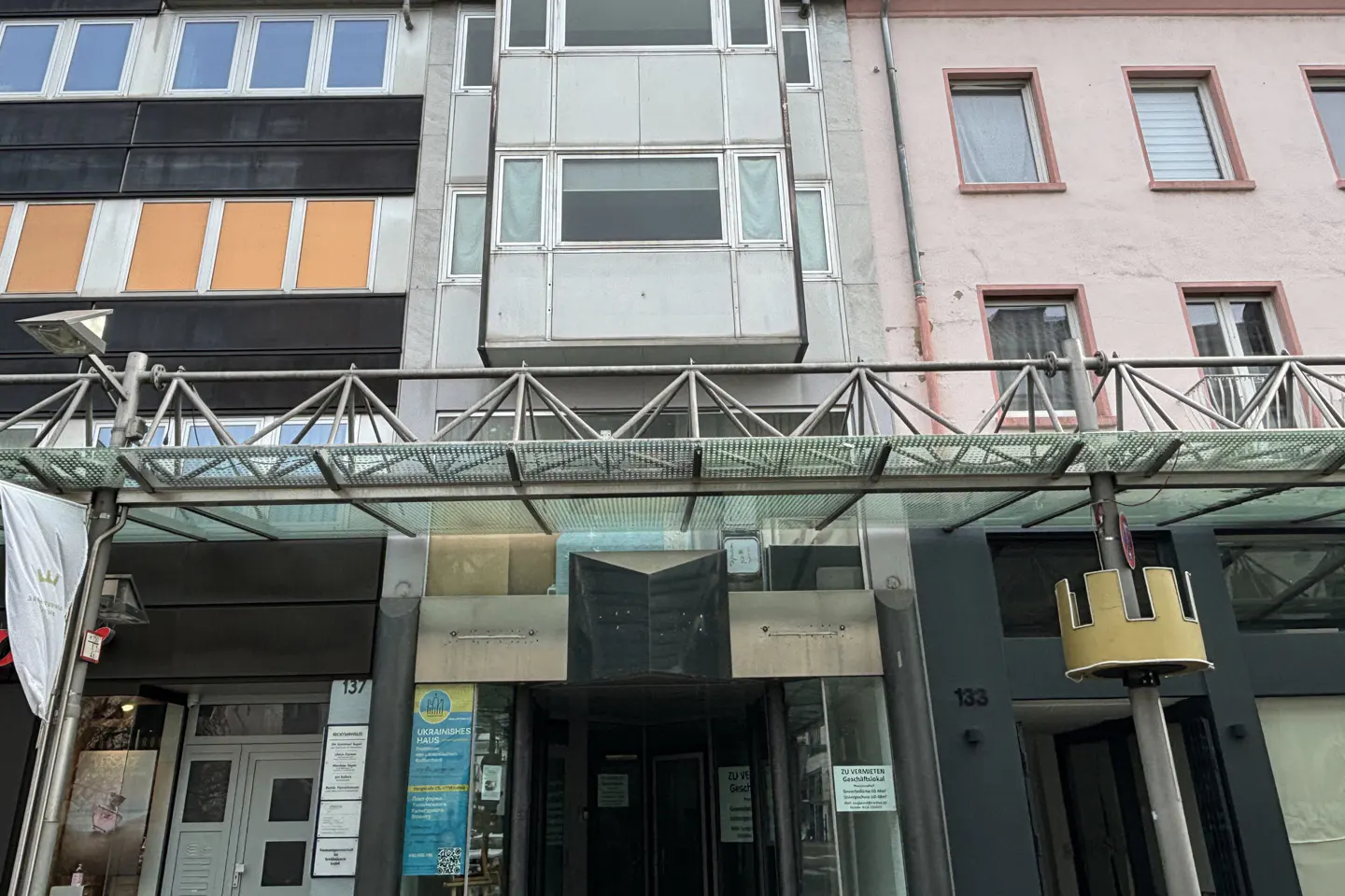 Street-level view of a modern building entrance with a glass canopy, flanked by commercial storefronts in a city.