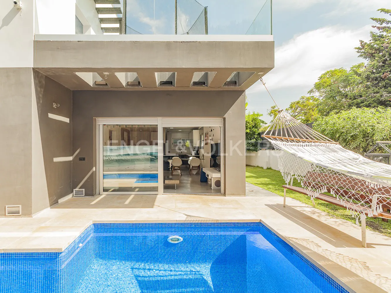 Exterior view of a modern house with a blue tiled pool, a white hammock, and sliding glass doors leading to the interior.