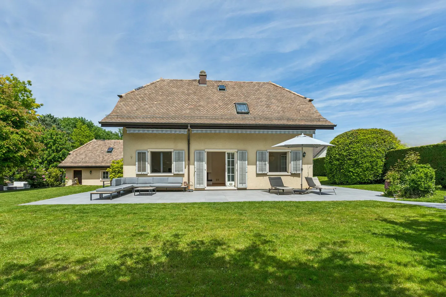 Back view of a tan house with a brown roof, patio furniture, and a green lawn under a blue sky.