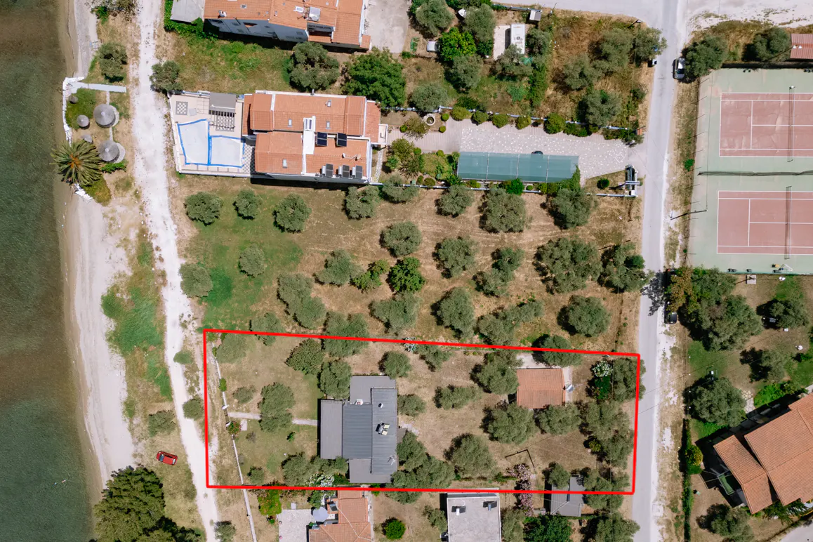 Aerial view of a property outlined in red, featuring a house, trees, and proximity to the beach and tennis courts.