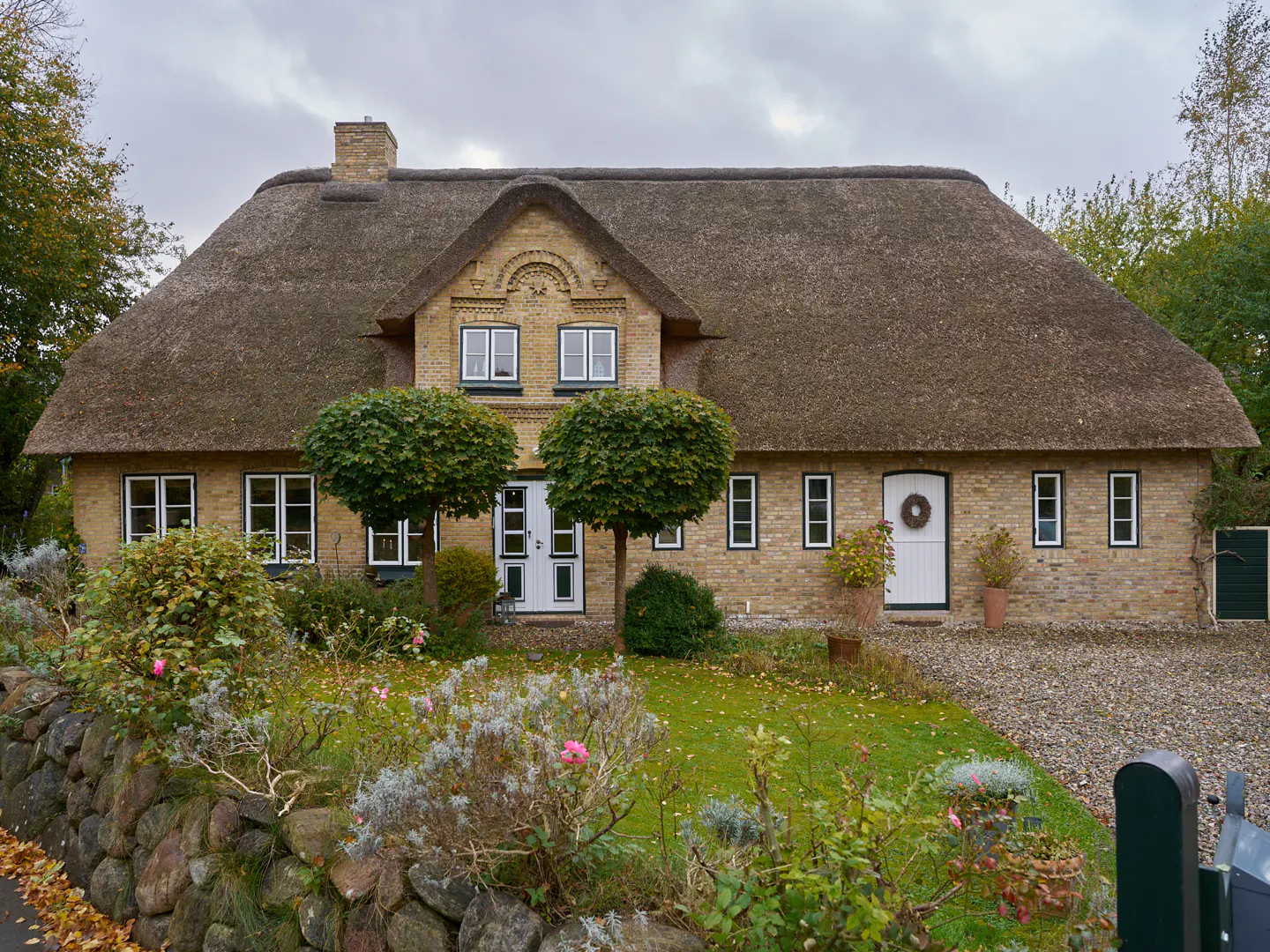 Two-story tan brick house with a thatched roof, white trim, and two white doors. A stone wall and garden are in the foreground.