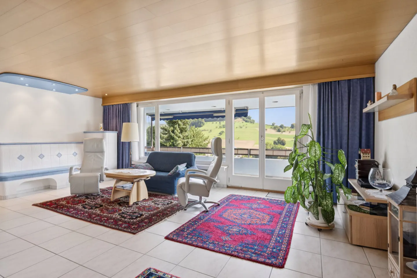 Bright living room with wood ceiling, white tile floor, blue sofa, and large windows overlooking green hills.