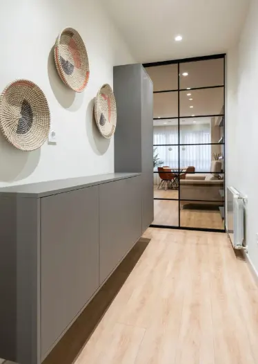 Hallway with light wood floors, gray cabinets, and decorative woven baskets on the wall. A black-framed glass door reveals a dining area.