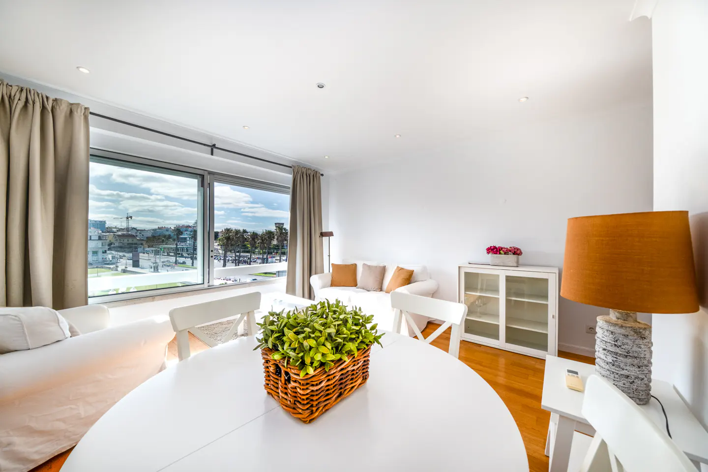 Bright living room with white walls, wood floors, and large window. A white table with a plant sits in the foreground. A white sofa is near the window.