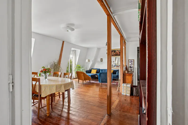 Open-plan living area with hardwood floors, a dining table, and a blue sofa set under slanted white walls.